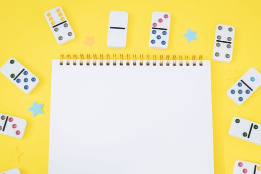White dominoes with colored dots are arranged in a circle around a blank notepad on a yellow background with copy space in the center, closeup flat lay. Summer board game concept.