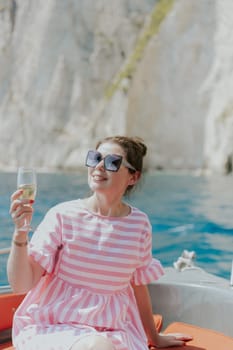 Portrait of one young Caucasian beautiful girl in sunglasses, a pink dress sits on the stern of a boat, holds a glass of champagne in her hand, looks to the side with a happy smile and enjoys sailing on the sea on a private boat on a sunny summer day, close-up side view.