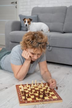 Mature Caucasian woman lies on the floor and plays chess. Jack Russell Terrier dog lying on the sofa