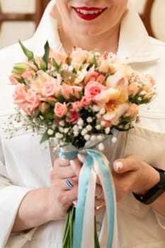 A woman is holding a bouquet of pink flowers with white accents. She is wearing a white jacket and has red lipstick on. The bouquet is tied with a blue ribbon