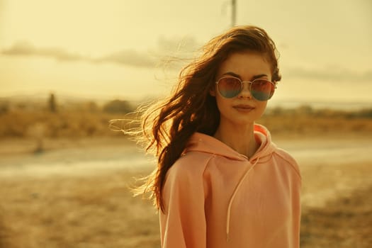 Stylish woman in pink hoodie and sunglasses poses confidently in vast field under clear blue sky