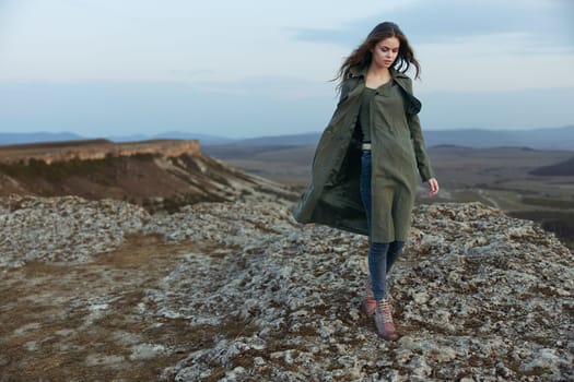confident woman in trench coat standing proudly atop hill with majestic mountains in background