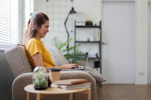 A young woman sits comfortably on a chair, using a laptop to browse social media in a bright, modern living room.