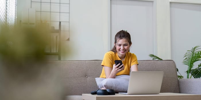 A young woman uses her smartphone and laptop in a modern living room, representing a social media home environment.