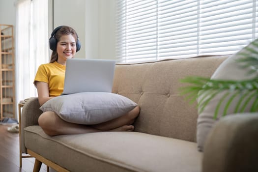 A young woman wearing headphones, sitting on a sofa, and enjoying music while using a laptop in a bright and modern living room.