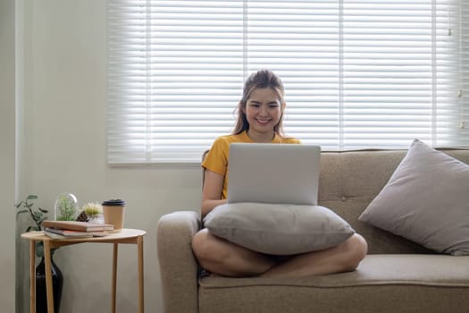 A young woman sits on a sofa at home, using a laptop to browse social media. The modern living room is bright with natural light.