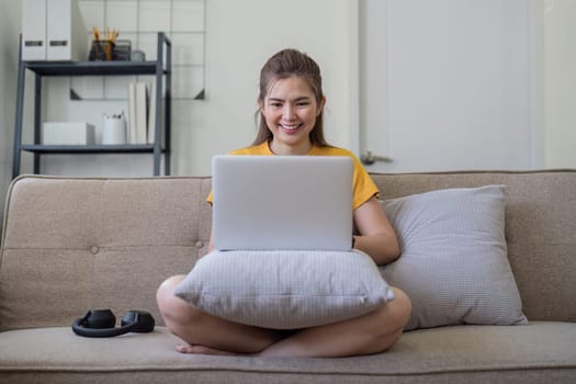 Young woman using laptop at home, enjoying social media, sitting on sofa in modern living room, smiling and relaxed.