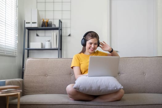 A young woman wearing headphones, sitting on a sofa, enjoying music on her laptop in a bright, modern living room.