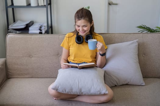 Young woman reading a book at home on a sofa, enjoying leisure time with a cup of coffee, wearing casual clothing and headphones.