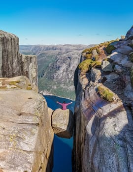 A lone hiker sits on a boulder overlooking a dramatic cliff face in Kjeragbolten Norway.