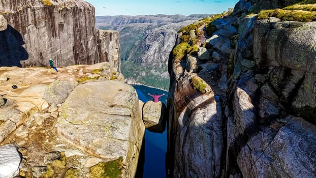 A hiker sits on the edge of Kjeragbolten, also known as the Pulpit Rock, in Norway, overlooking a deep valley and a stunning fjord landscape.