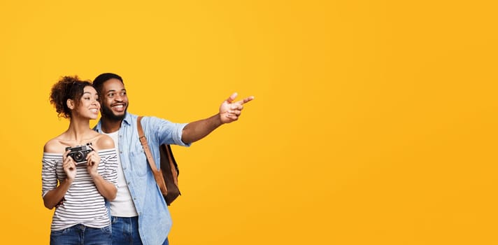 African American man and woman are pointing at something off-camera with their fingers extended, standing against a vibrant yellow background, going vacation, copy space