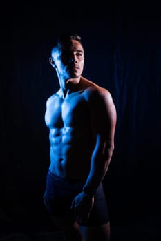 A man with a bare chest is posing against a dark backdrop, showing off his muscular physique. His head, hands, stomach, arms, shoulders, legs, and neck are visible in the flash photography