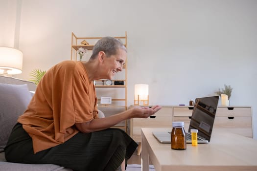 Elderly woman maintaining her health at home, engaging in an online consultation via laptop, with medication bottles on the table, in a modern living room setting.