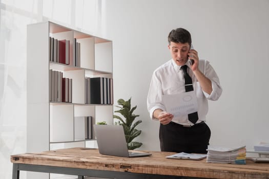 A young businessman in a modern office, looking disappointed while talking on the phone and holding documents.