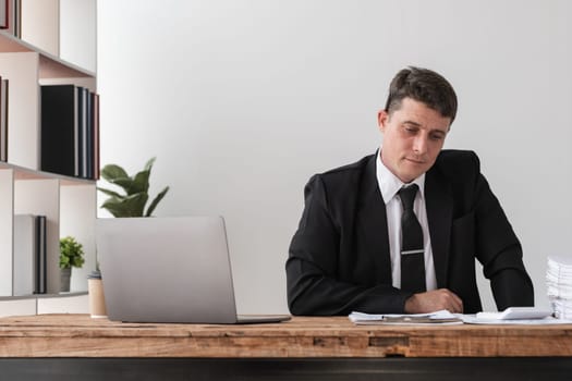 A young businessman in a suit looks disappointed while working at his desk in a modern office, surrounded by a laptop, documents, and shelves.