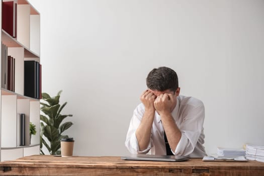 Young businessman sitting at a desk with head in hands, showing disappointment and frustration at work. Office environment with modern decor.