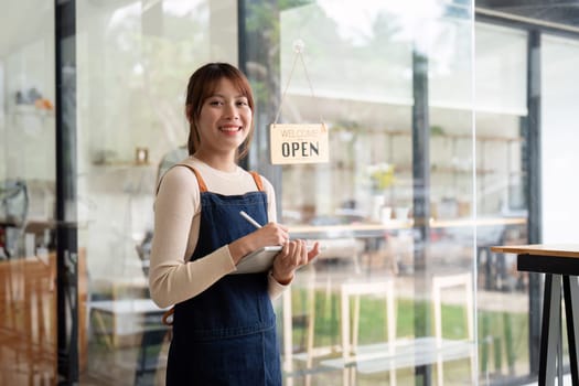 A young woman cafe owner smiling and holding a notepad in her modern coffee shop, ready to welcome customers with an Open sign on the door.