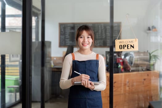 A young woman cafe owner in an apron, holding a notebook, standing in her modern coffee shop with an open sign, representing small business entrepreneurship.