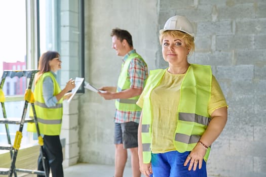 Female designer managing finishing decoration in safety helmet vest looking at camera with team workers in background in cement room of office. Industrial workers construction repair work concept