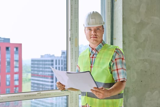 Portrait of young male industrial worker in safety vest helmet looking at camera with working engineering architectural documents on construction. Construction repair service development work concept