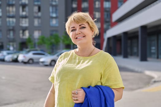 Portrait happy elegant mature woman 50 years old outdoor, urban background. Smiling beautiful blonde female in yellow looking at camera in city. Age, lifestyle, beauty, fashion concept