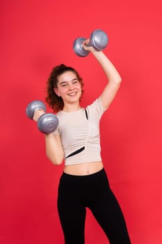 Teenage sportive girl exercises with dumbbells to develop muscles isolated on a red background.