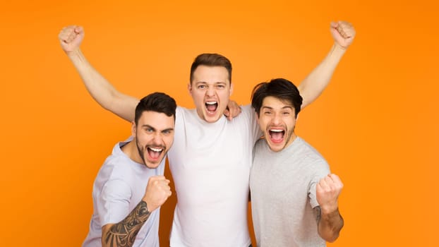 Three young men are exuberantly celebrating, arms raised in triumph, against a vibrant orange background