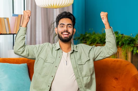 Happy Indian man shouting, celebrating success, winning, goal achievement good news, lottery luck, victory, watching TV championship at modern home apartment. Arabian guy in living room sits on couch