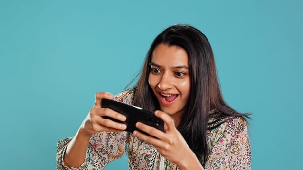 Happy woman playing videogames on smartphone, celebrating after defeating foes, doing clenching hand gesture. Player swiping phone screen, thrilled about winning game, studio background, camera B