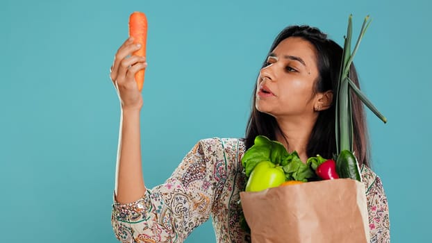 Woman with paper bag in hands filled with vegetables checking carrot for impurities. Conscious living customer with purchased groceries to be used as cooking ingredients, studio background, camera B