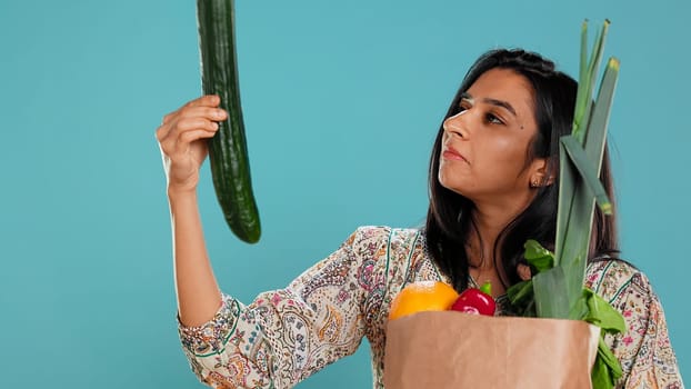 Woman with paper bag with vegetables testing quality, looking at cucumber, isolated over studio background. Person inspecting groceries after buying them from zero waste shop, camera B