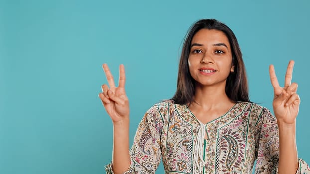 Jolly indian person doing peace victory hand sign gesture, studio backdrop. Portrait of upbeat woman, feeling confident, celebrating win, isolated over studio background, camera B