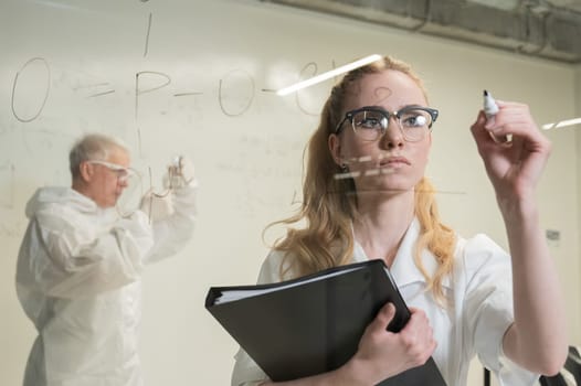 A woman chemist writes a formula on glass. An elderly Caucasian man in a protective suit is doing tests