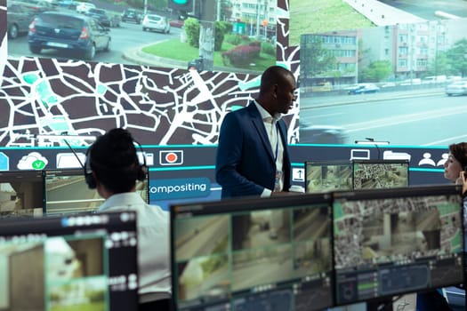 African american supervisor managing his team in control center at shipping agency headquarters, overseeing shipping activities to ensure order and parcel delivery. Working with satellite maps.