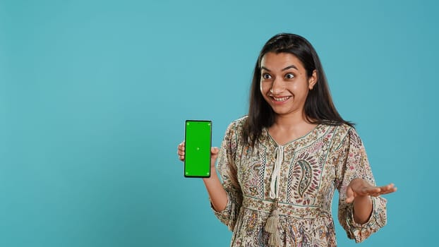 Tech content creator doing influencer marketing using green screen smartphone, isolated over studio background. Indian woman holding empty copy space mockup phone, doing review, camera A