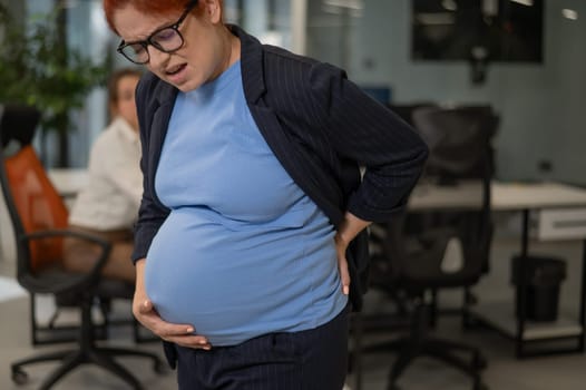 A pregnant woman suffers from pain and holds her stomach while standing in the middle of the office