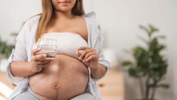 A pregnant woman sits comfortably in a modern living room, holding a glass of water, surrounded by cozy decor and indoor plants, embracing motherhood.