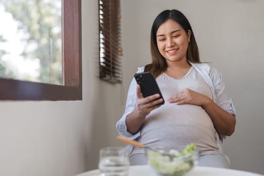 A pregnant woman sits comfortably in a modern living room, using her smartphone and enjoying a healthy snack. Natural light fills the cozy home environment.