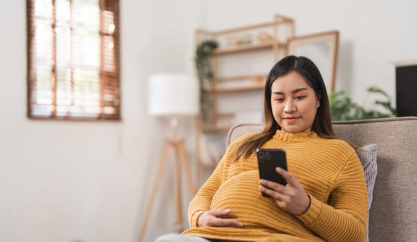 A pregnant woman sits comfortably in a modern living room, using her smartphone. The cozy home interior is filled with natural light, creating a peaceful and serene atmosphere.