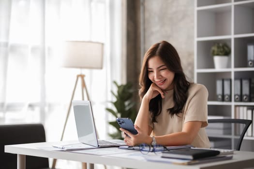 Young female accountant working from home, using a laptop and smartphone in a modern home office with natural light and minimalist decor.