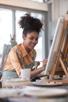 A young African woman sits in a bright studio, painting on a canvas. She is wearing an apron and casual clothing, surrounded by art supplies and natural light.