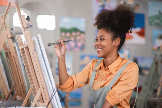 Young African woman painting on canvas in a vibrant art studio, wearing an apron and smiling, surrounded by colorful artwork.