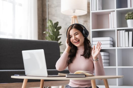 Young woman working in a modern living room, using a laptop and headphones, smiling and waving, surrounded by books and cozy decor.