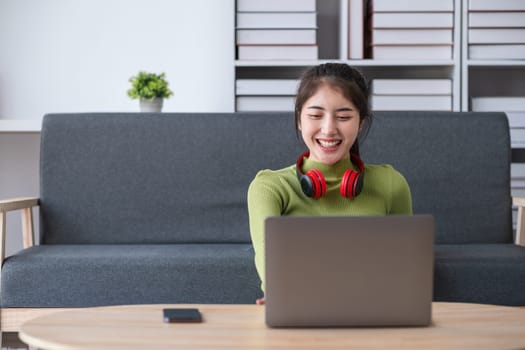 Young woman sitting in a modern living room, working on a laptop with headphones, smiling and engaged in remote work or study.