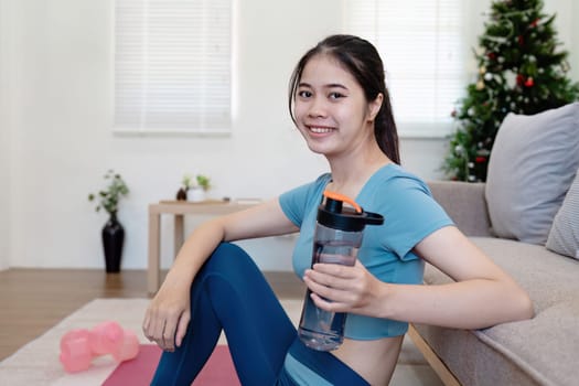 Young woman in activewear exercising at home, holding a water bottle, with yoga mat and dumbbells in a modern living room.