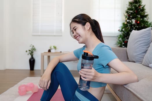 A young woman in activewear exercising at home, sitting on a yoga mat with dumbbells and a water bottle, smiling in a bright, modern living room.