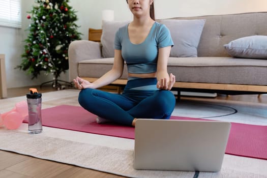 Woman doing yoga on a mat in a cozy living room, with a laptop and exercise gear nearby, and a Christmas tree in the background.