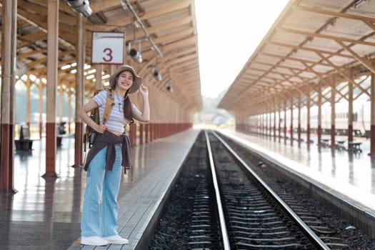 A young female traveler standing on a train station platform, ready for an adventure. She is wearing a backpack and hat, smiling with excitement as sunlight streams through the wooden roof structure.