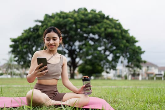 Young woman practicing yoga outdoors, sitting on a pink mat, holding a smartphone and water bottle, promoting wellness and mindfulness in a green park.